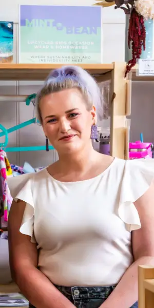 Jac Travers smiling and standing infront of a sign for Mint & Bean (upcycled clothing and homewares)