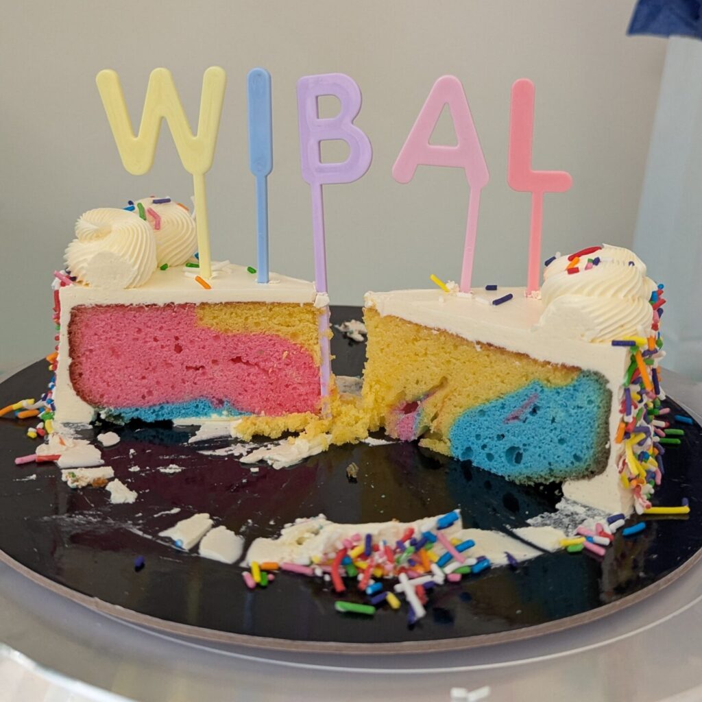 A cut up rainbow cake with white icing. The cake topper has the acronym WIBAL on the top in pastel rainbow.