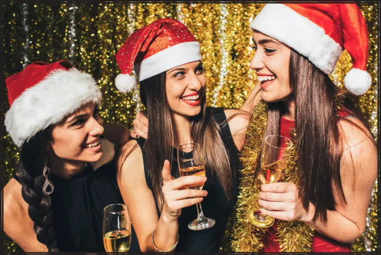 Three brunettes with Santa hats on, are drinking and smiling at each other during a Christmas party