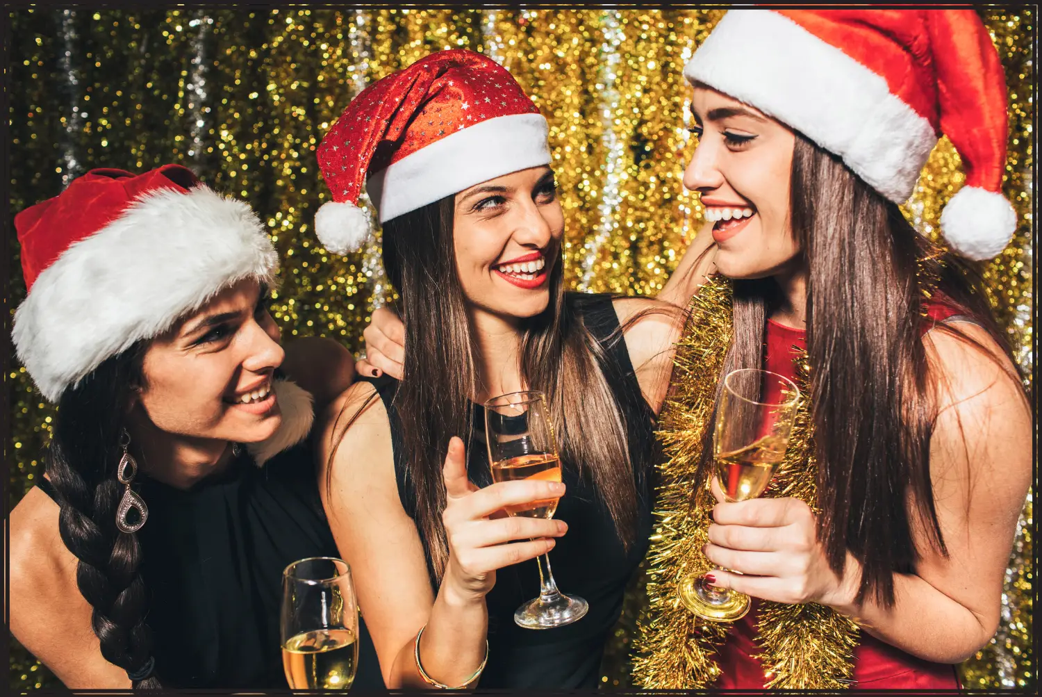 Three brunettes with Santa hats on, are drinking and smiling at each other during a Christmas party